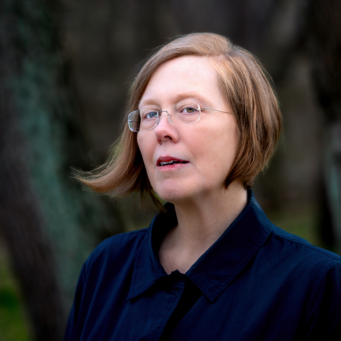 Woman with chin-length strawberry blonde hair wearing a blue jacket in a dark forest
