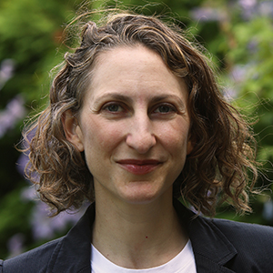 A woman with curly dark blonde hair who is wearing a black blazer and white undershirt.