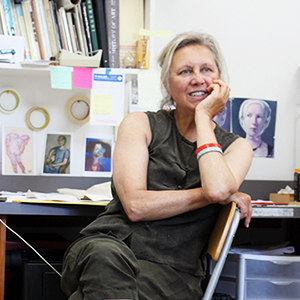 Woman in a gray outfit looking slightly away from the camera with her chin resting in her hand sitting in front of her desk with various prints on the wall.