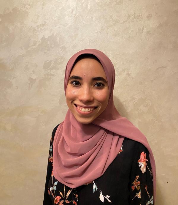 a woman smiling with a pink headdress and black floral dress in front of a cream colored textured wall