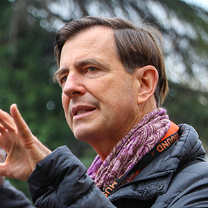 headshot of a man with brown hair wearing a multicolor scarf and gesturing with his hand