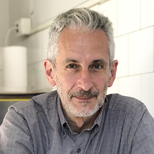 Man with beard add gray patterned shirt sitting in a studio with tiled walls.