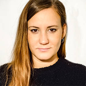 A person with brown hair and a black shirt smiling in front of a beige wall.