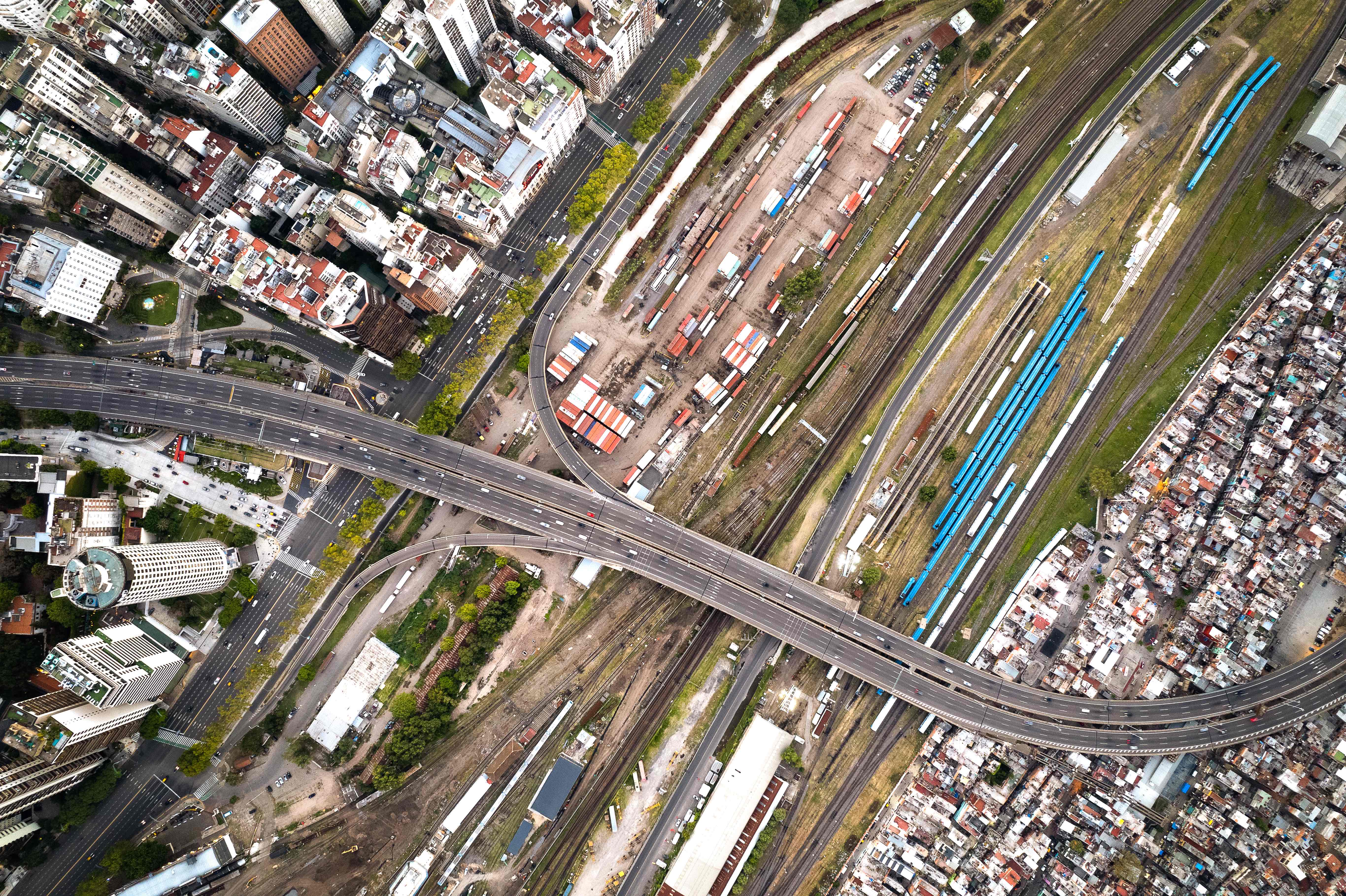 an aerial view of an urban area with a complex overpass system. Multiple lanes of traffic on the overpass intersect at various points, with cars visible on the roads. Below the overpass, there are train tracks with several blue train cars stationed along them. The area surrounding the overpass and train tracks appears densely packed with buildings of varying sizes, shapes, and colors, indicative of a crowded city environment.