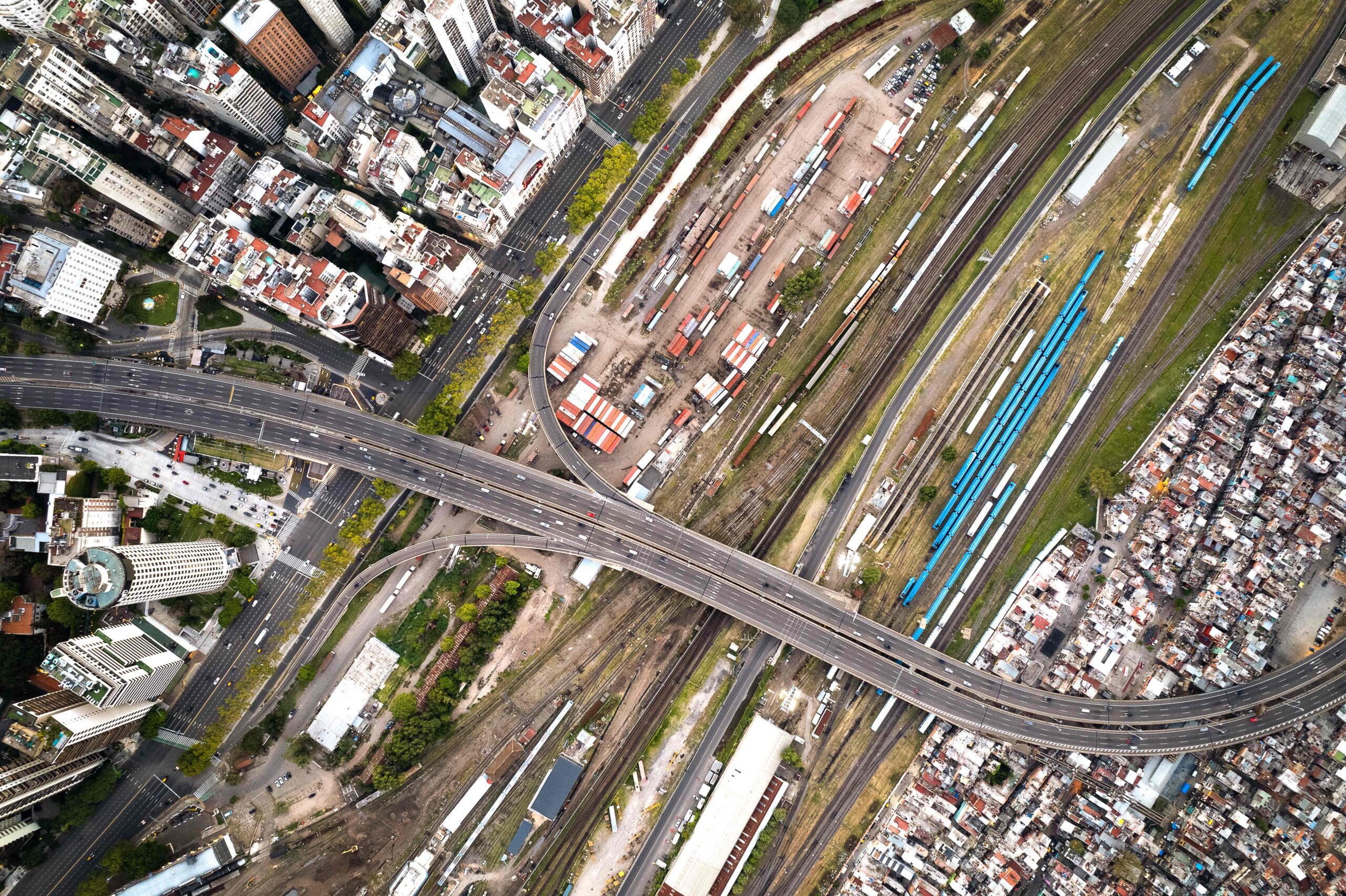 an aerial view of an urban area with a complex overpass system. Multiple lanes of traffic on the overpass intersect at various points, with cars visible on the roads. Below the overpass, there are train tracks with several blue train cars stationed along them. The area surrounding the overpass and train tracks appears densely packed with buildings of varying sizes, shapes, and colors, indicative of a crowded city environment.