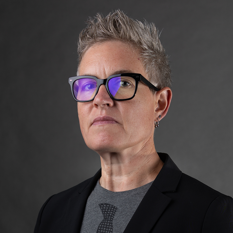 A woman with short spiked hair wearing glasses and a black suit jacket with a grey shirt underneath.