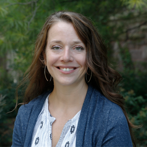 A woman with long brown hair wearing earrings and a blue sweater standing outside smiling.