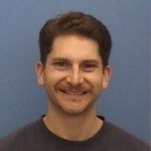 A person smiling with short dark hair wearing a dark blue shirt