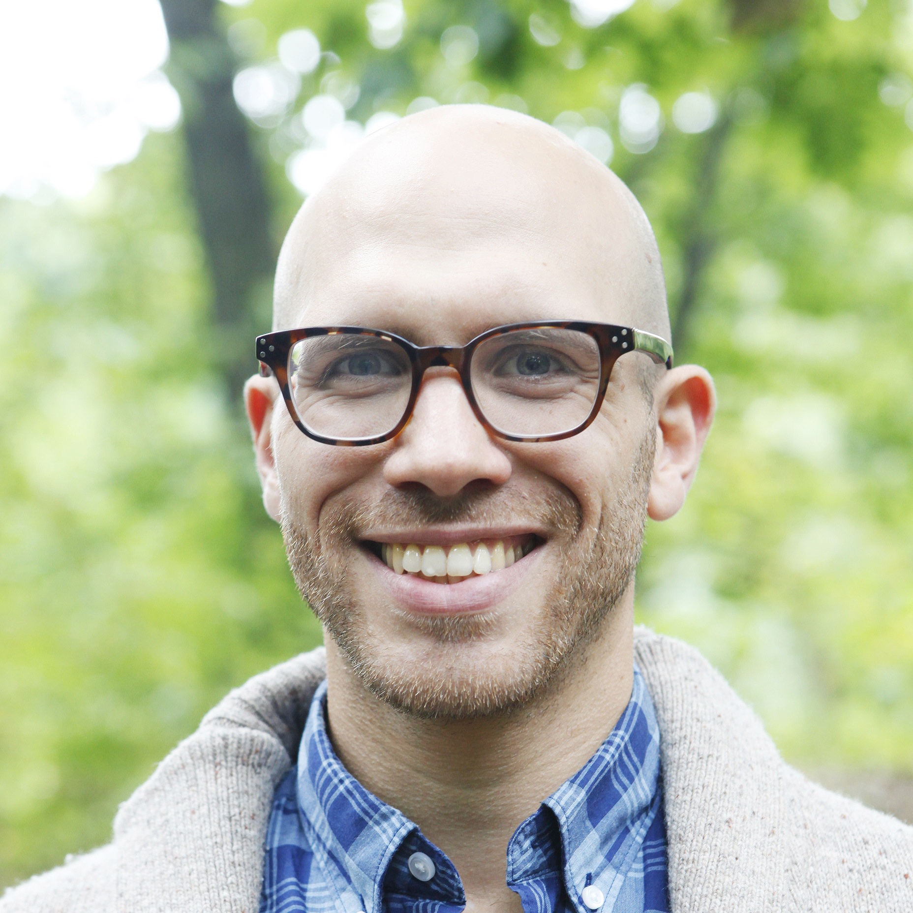 headshot of a man with tortoise shell glasses wearing a blue plaid shirt and a tan sweater