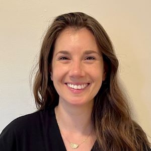 A person smiling with long brown hair wearing a black top in front of a white background