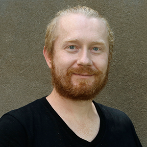 A person with a black vneck and a red beard standing in front of a concrete wall.