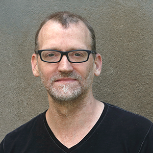 A man with a black t-shirt wearing black framed glasses standing in front of a concrete wall.