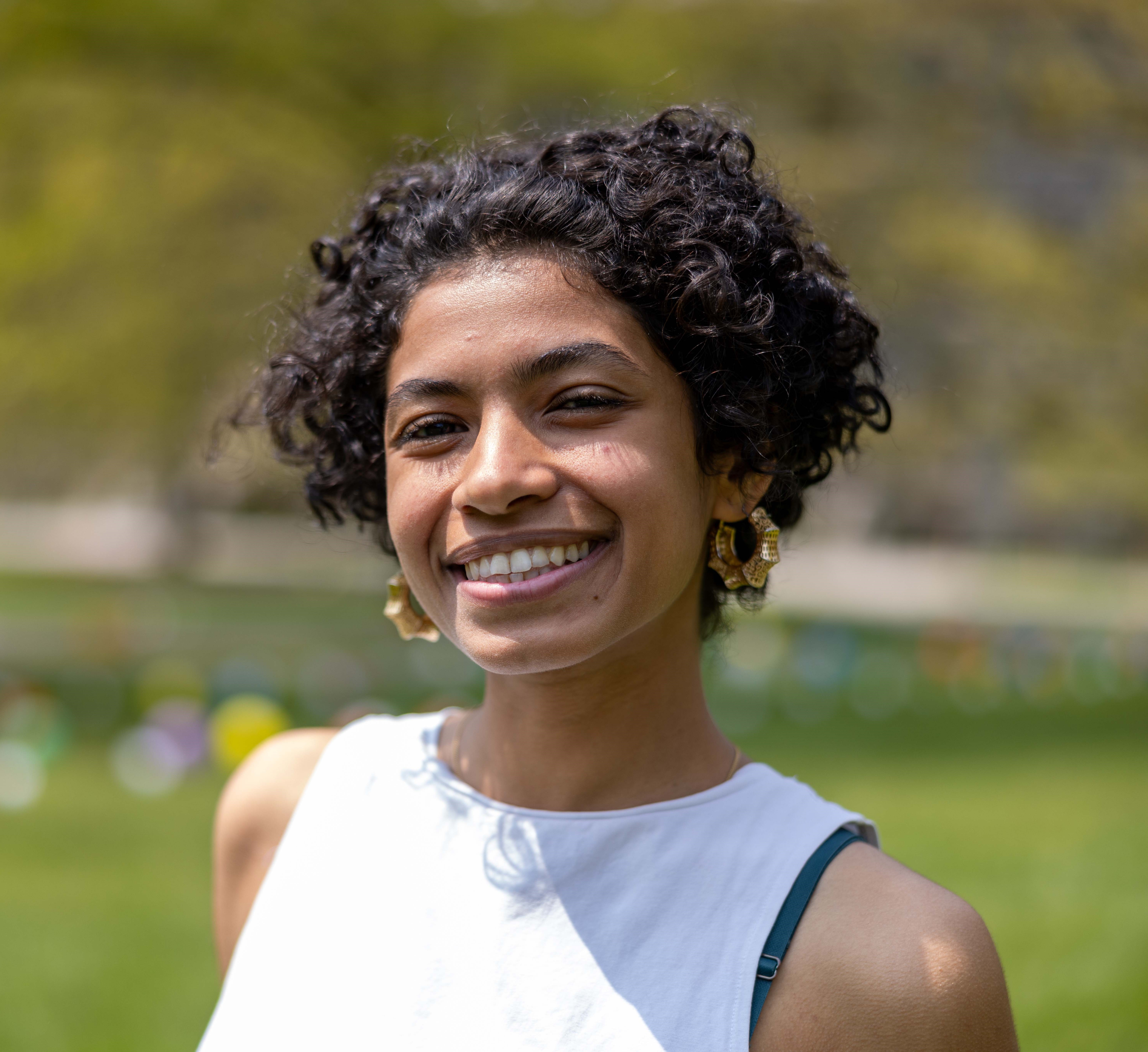 A person smiling with a curly hair, a white tank top shirt and gold earrings.
