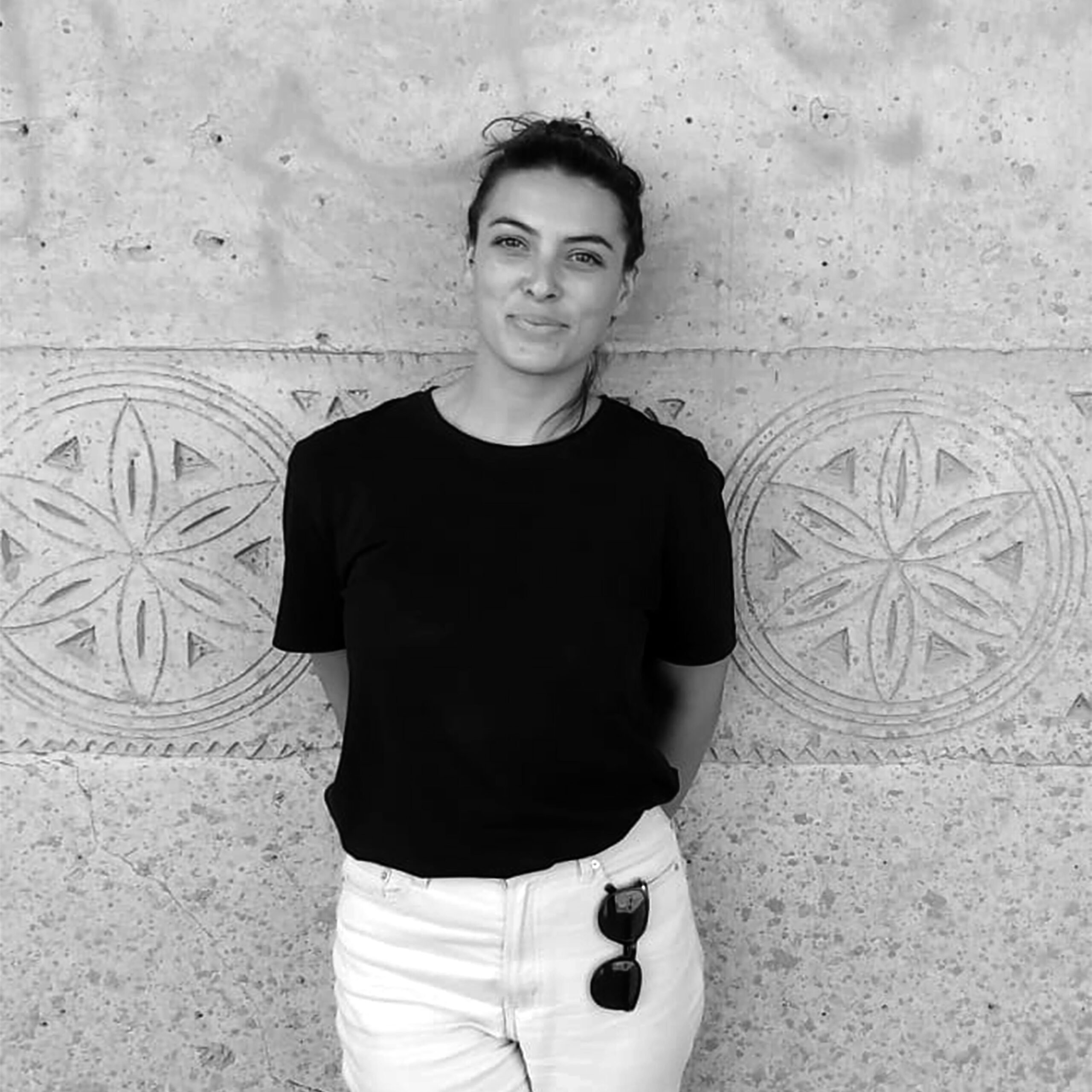 A woman in a black t-shirt and white jeans standing against a stone wall.