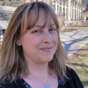A person with long blond hair wearing a black top and silver necklace standing outside in front of a building
