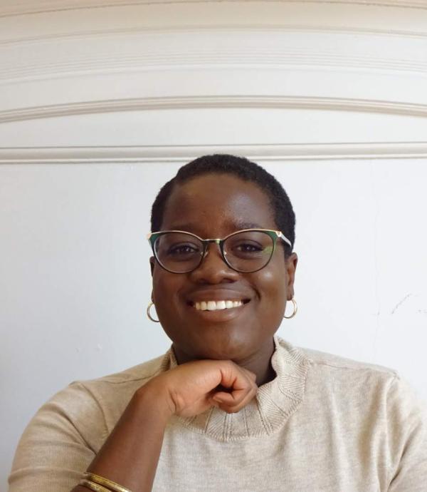 a person with glasses, gold hoop earrings, and gold bracelets wearing a cream colored shirt in front of a white wall