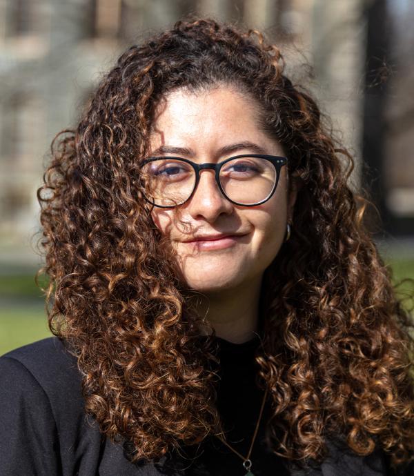 a young woman wearing black framed glasses with long curly brown hair standing outside wearing a black shirt and a gold necklace.