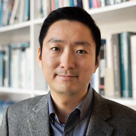 A man in grey jacket and navy blue shirt smiling in front of book shelf