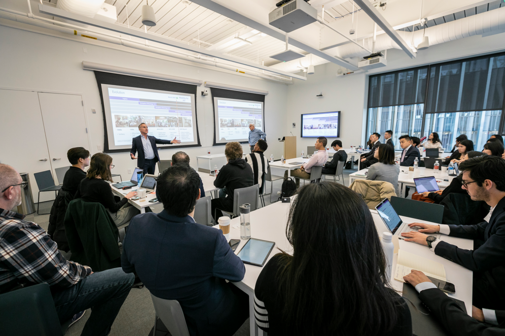 Two men standing in front of a group of students delivering a lecture.