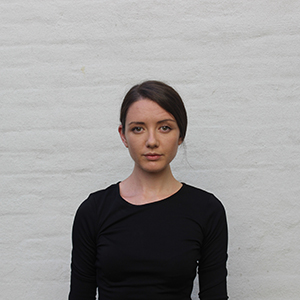 A person with dark brown hair wearing a long sleeve black shirt. Grey brick wall..