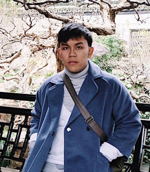a young man with short dark hair wearing a turtle neck and a blue jacket with a black strap of a bag draped over one shoulder