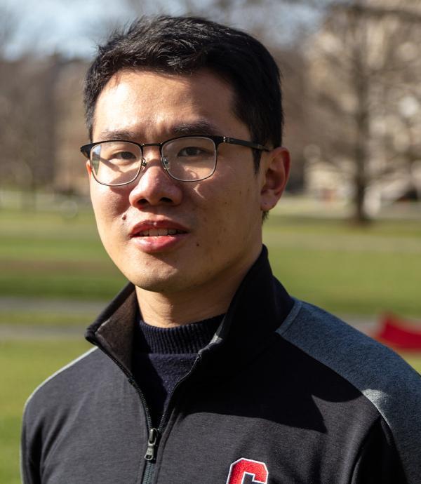 a young man with short black hair standing outside wearing black framed glasses and a quarter zip sweater with an embroidered Cornell 