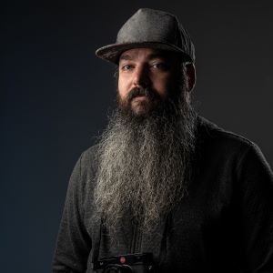A person with a long dark beard wearing a grey cap and grey sweater holding a camera in front of a dark background
