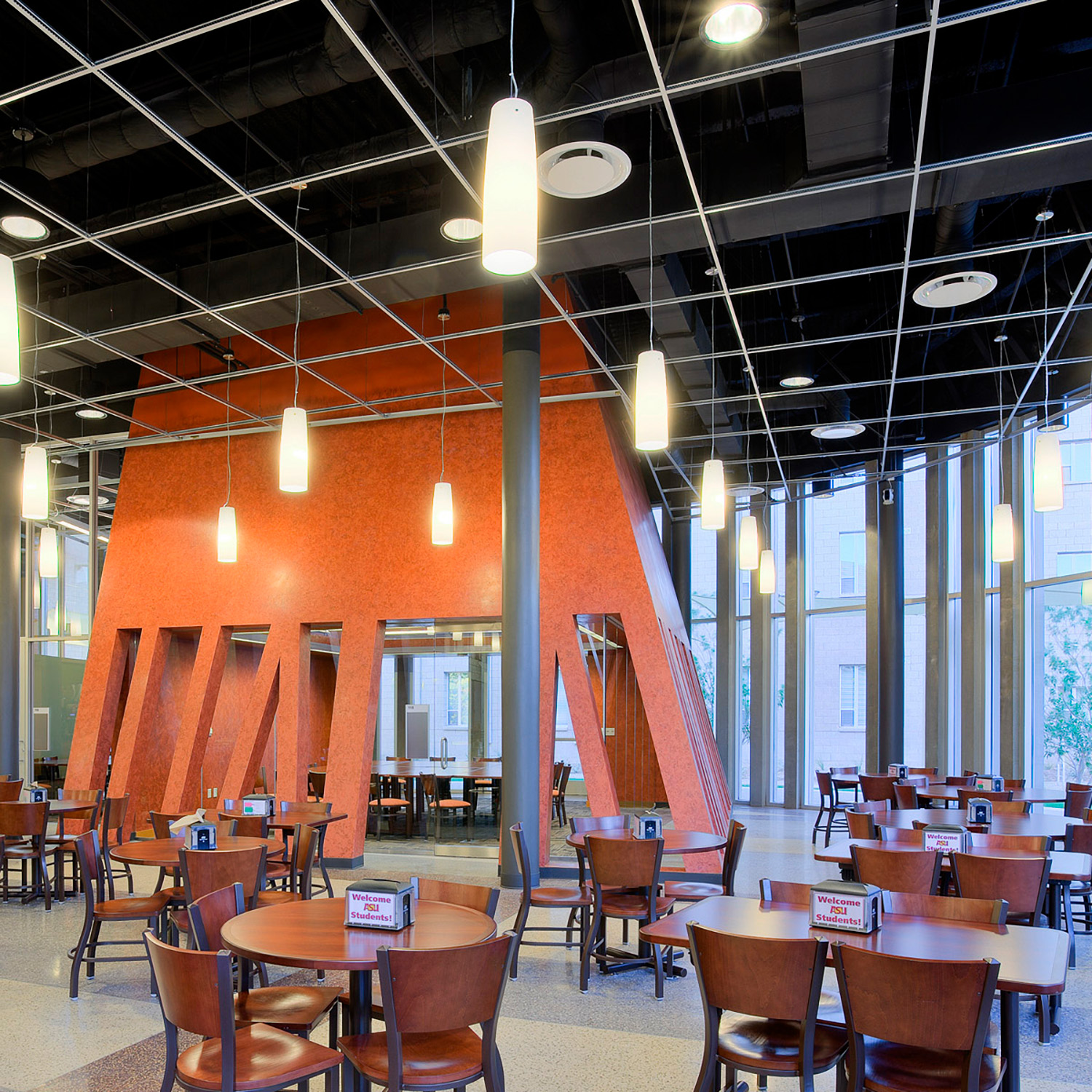 A dining hall with tables surrounding an orange structure with tall rectangular windows showing a large dining table inside a private dining room.