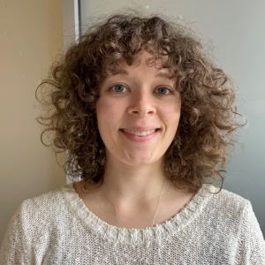 A person smiling with long curly brown hair wearing a white sweater