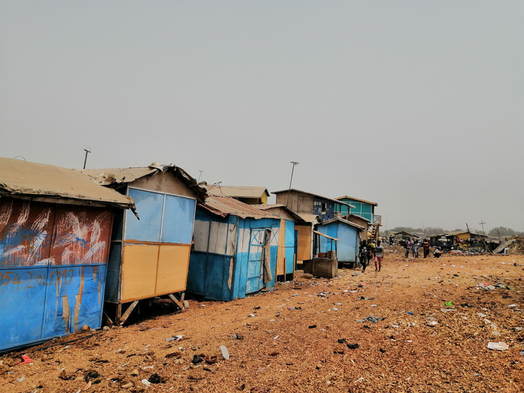 A row of shelters built from colorful sheets of metal with tan rooves extends into the distance. The rocky tan ground has trash scattered across it.