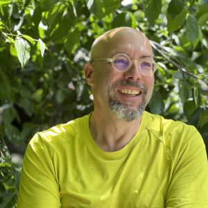 Smiling man with salt and pepper facial hair wearing round acrylic glasses and a citron colored tshirt