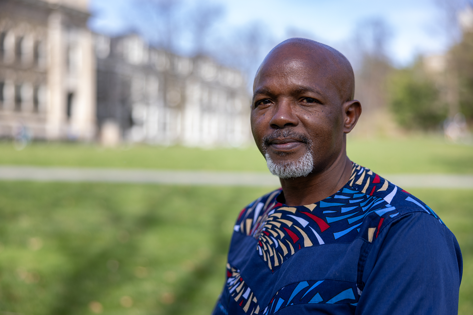 Dark skinned man with grey goatee wearing a blue embroidered shirt and standing on a green lawn in front of a grey stone building.
