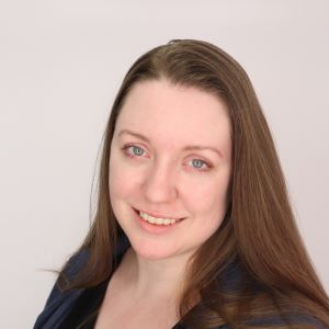 Person with long light brown hair parted to the side. They are standing in front of a white backdrop and are smiling at the camera. They have blue eyes and are wearing a blue shirt.