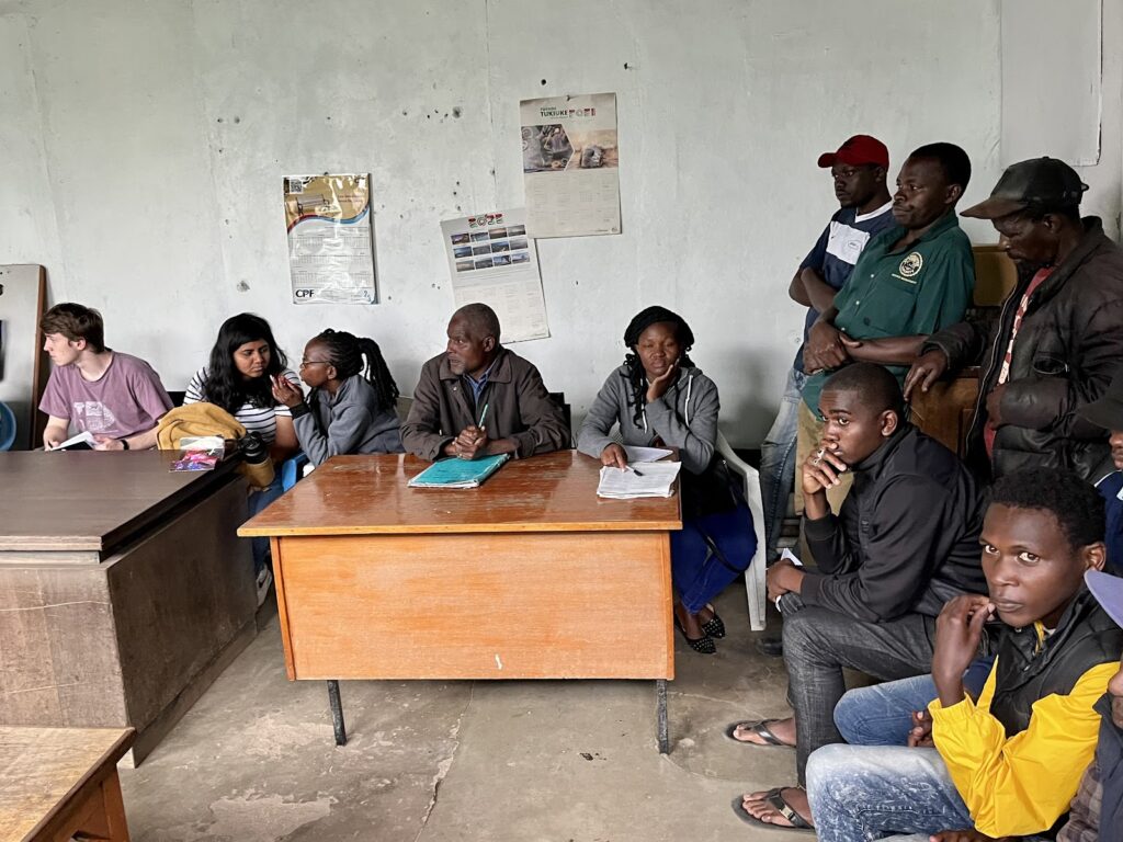 a group of people inside a simple room with gray walls, engaged in what appears to be a meeting. Across the room, a mix of men and women are standing or seated against the wall, attentively observing the interaction.