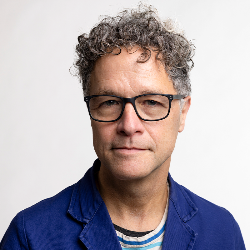 A man with curly hair wearing glasses and a striped shirt and blue jacket.