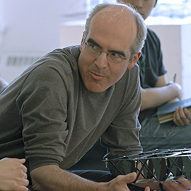man with glasses and gray hair leaning on a table talking to someone out of the frame