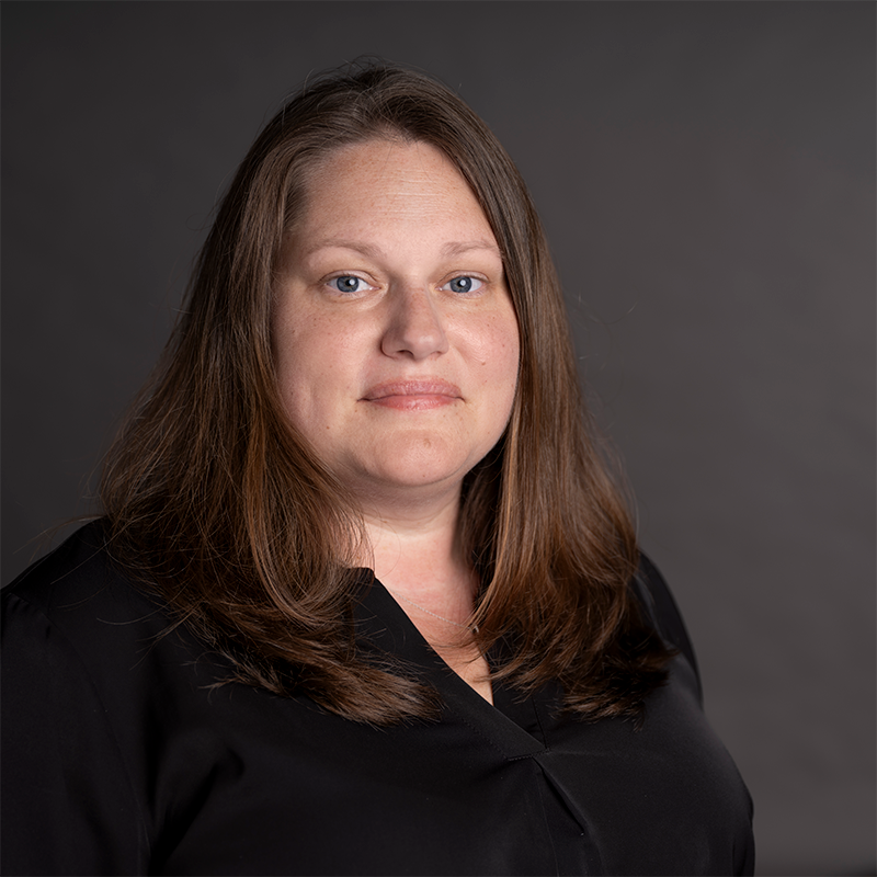 A person with long dark hair and blue eyes smiling in front of a green grey wall