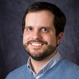 headshot of man with beard in blue sweater with dark gray back drop