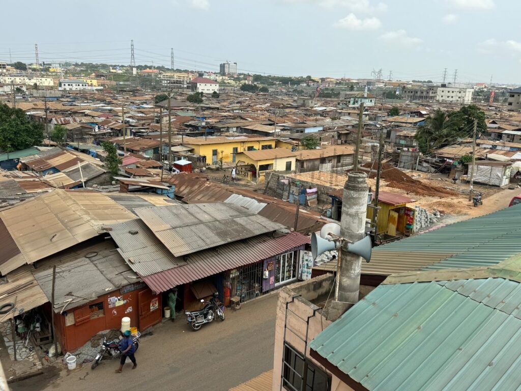 A view from a few stories up of a dense cluster of buildings made up of metal sheeting and metal rooves. A man walks with a motorcycle on the side of the road in the bottom of the photo.