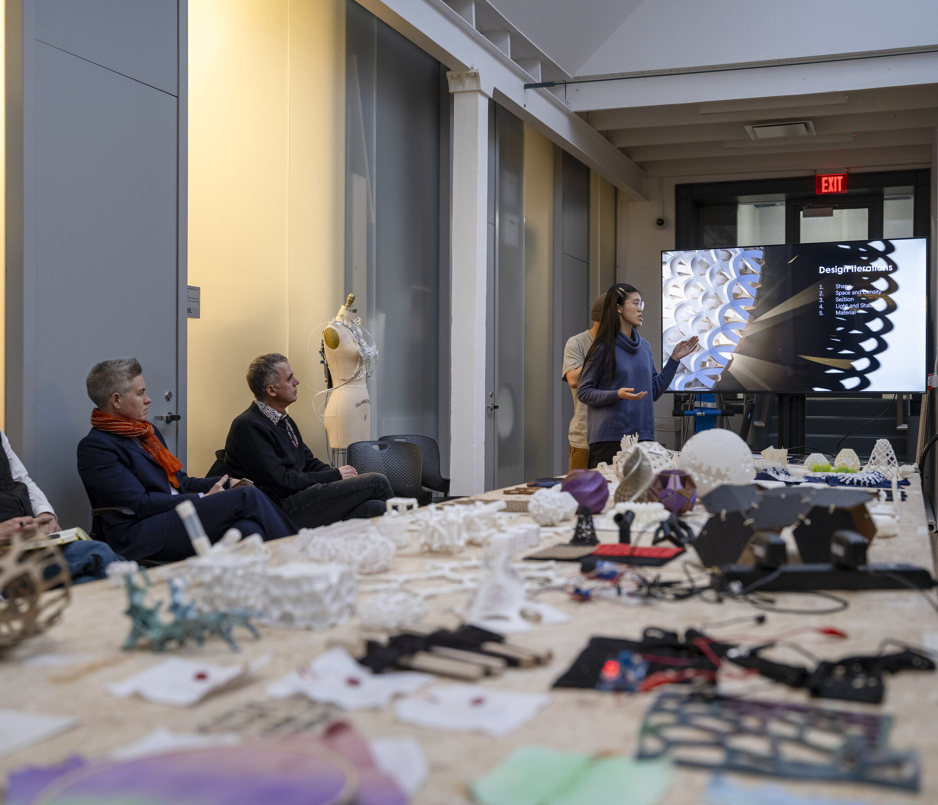 a person presenting a presentation on a large screen in front of people with 3d printed parts resting on a table