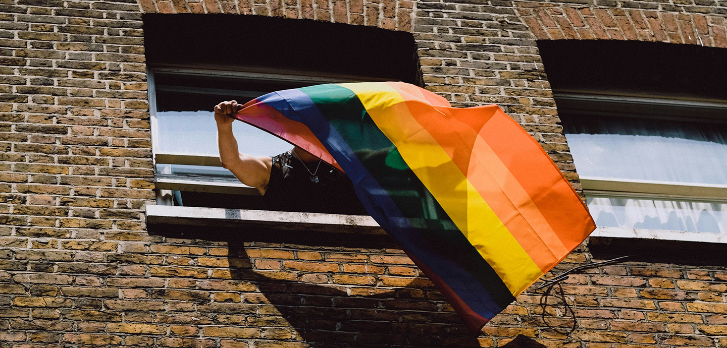 Man holding a flag of different strips of color from window of brick building