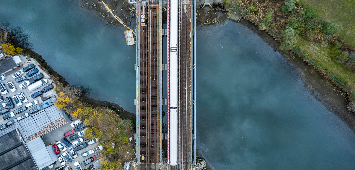 Aerial view of a train bridge over a river. Clouds are reflected in the water. In the upper right corner is the edge of land with green grass and trees. In the bottom left corner is land with some trees and about twenty cars and an industrial building.