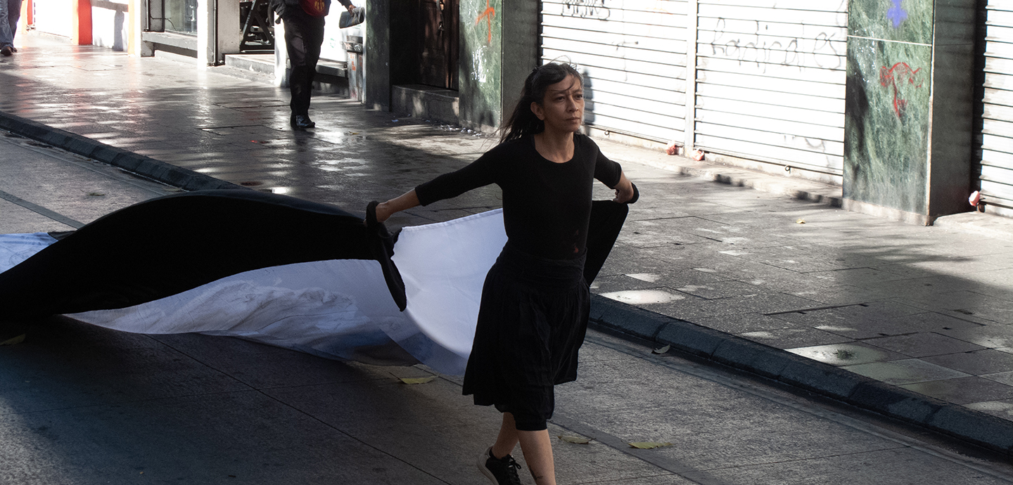 A petite woman with dark hair and wearing a black dress passes a graffitied building as she walks down a street