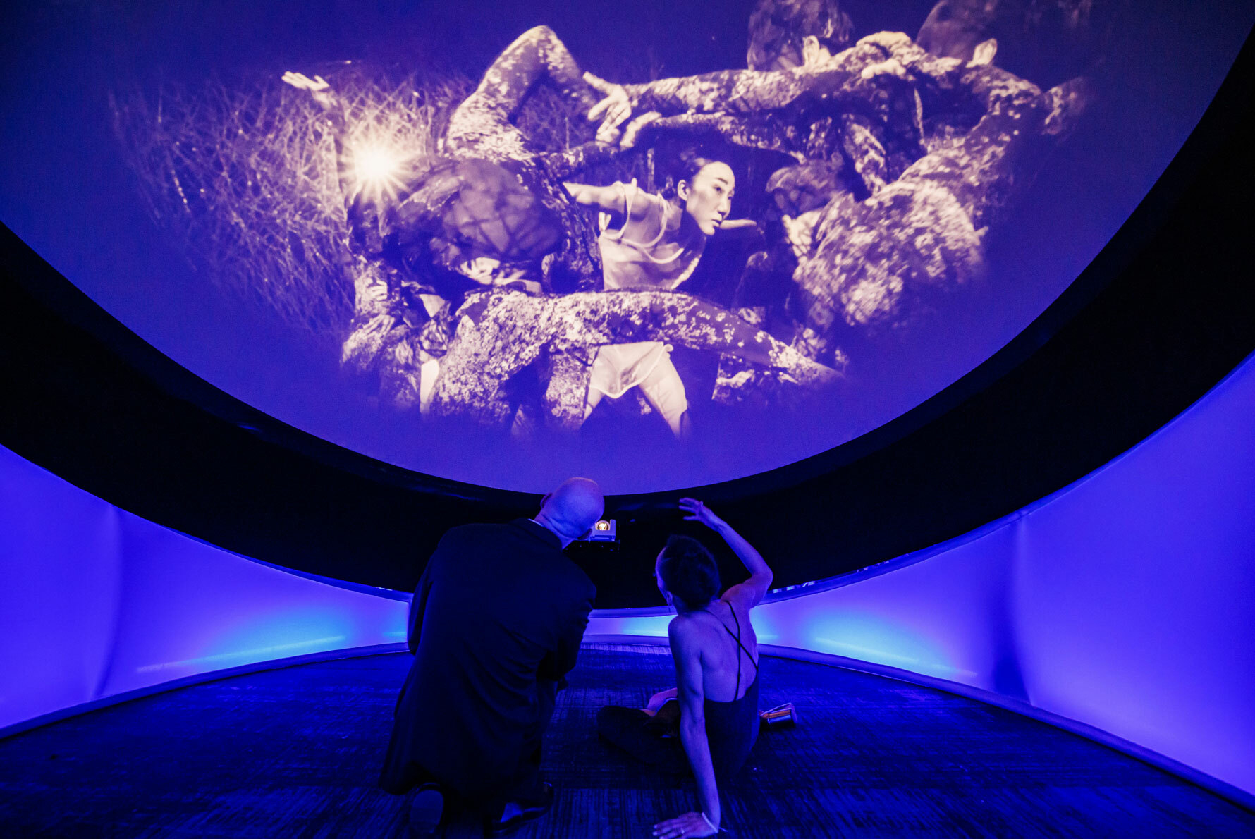 A man and a woman sit on a floor in a dimly lit circular room, cast with a purple glow, and watch a performance on a round screen