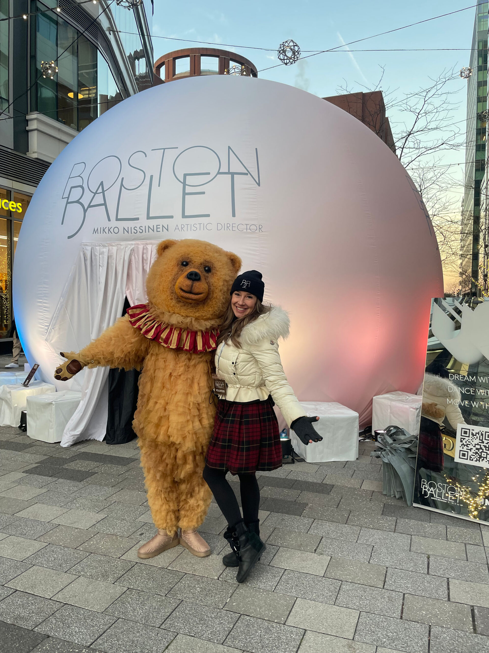 A woman and a person in a bear costume pose outside in front of a large white sphere with the words Boston Ballet printed on it