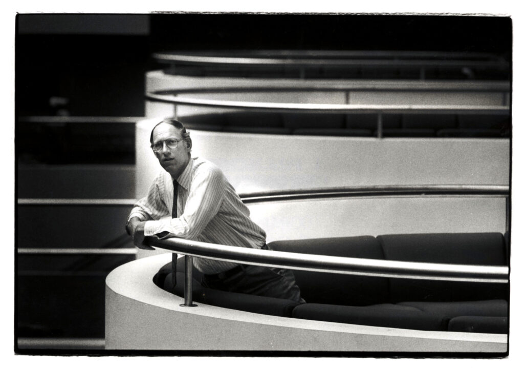 Black and white photo of a man in professional attire leaning against a balcony railing.