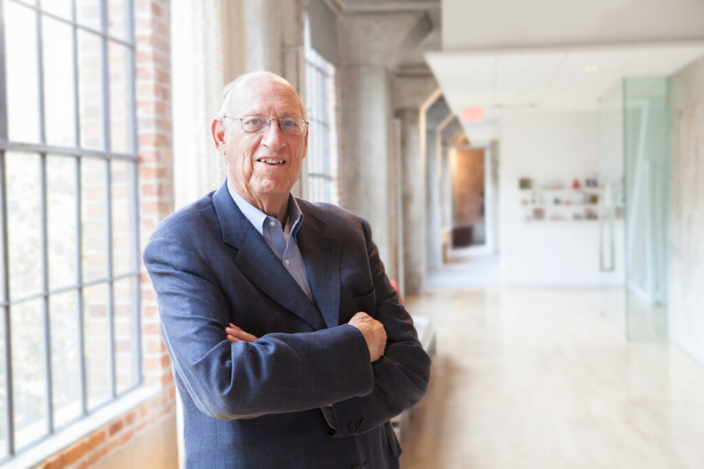 Portrait of an elderly man dressed in a navy blue suit jacket over a light blue button-up shirt, posing inside a well-lit hallway.