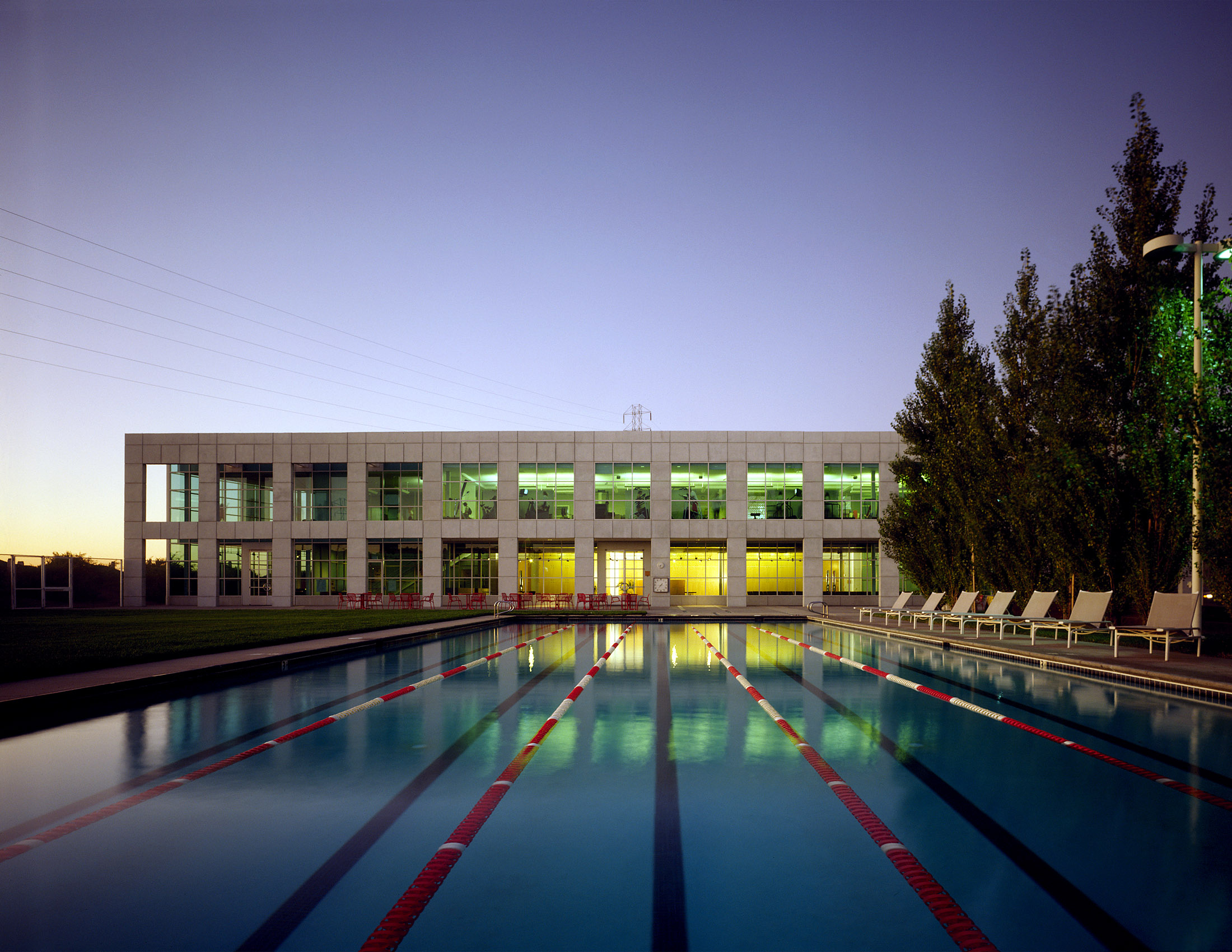 A long, multilane pool leads toward a two-story building lined with two rows of large windows lit up against a purple dusk sky.