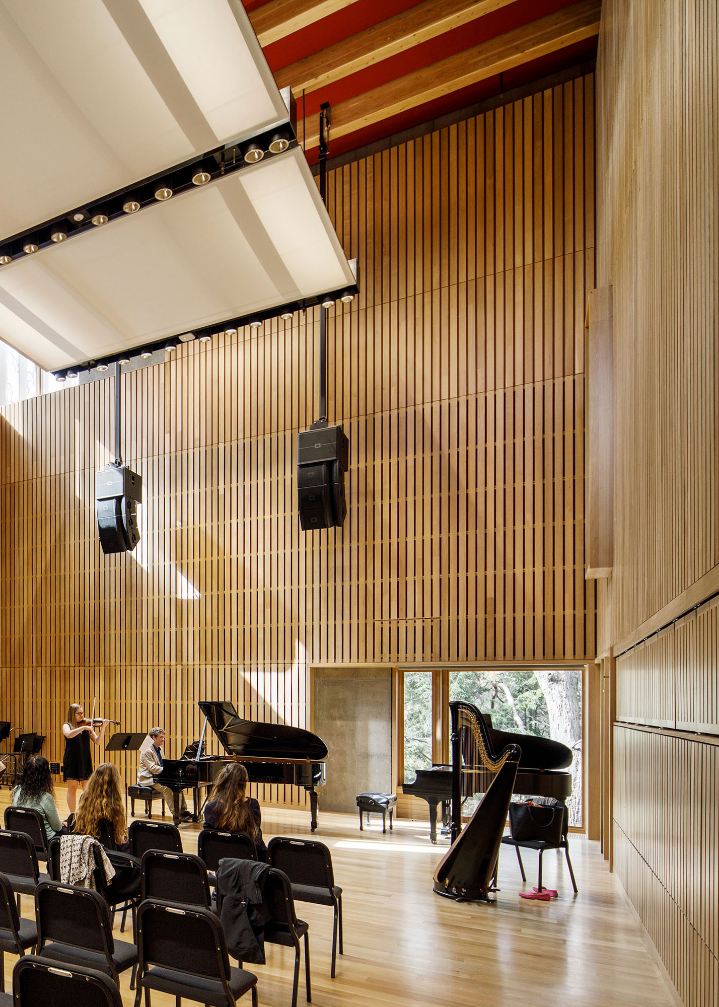 Three female students watch another student and a professor play instruments in a rehearsal hall.