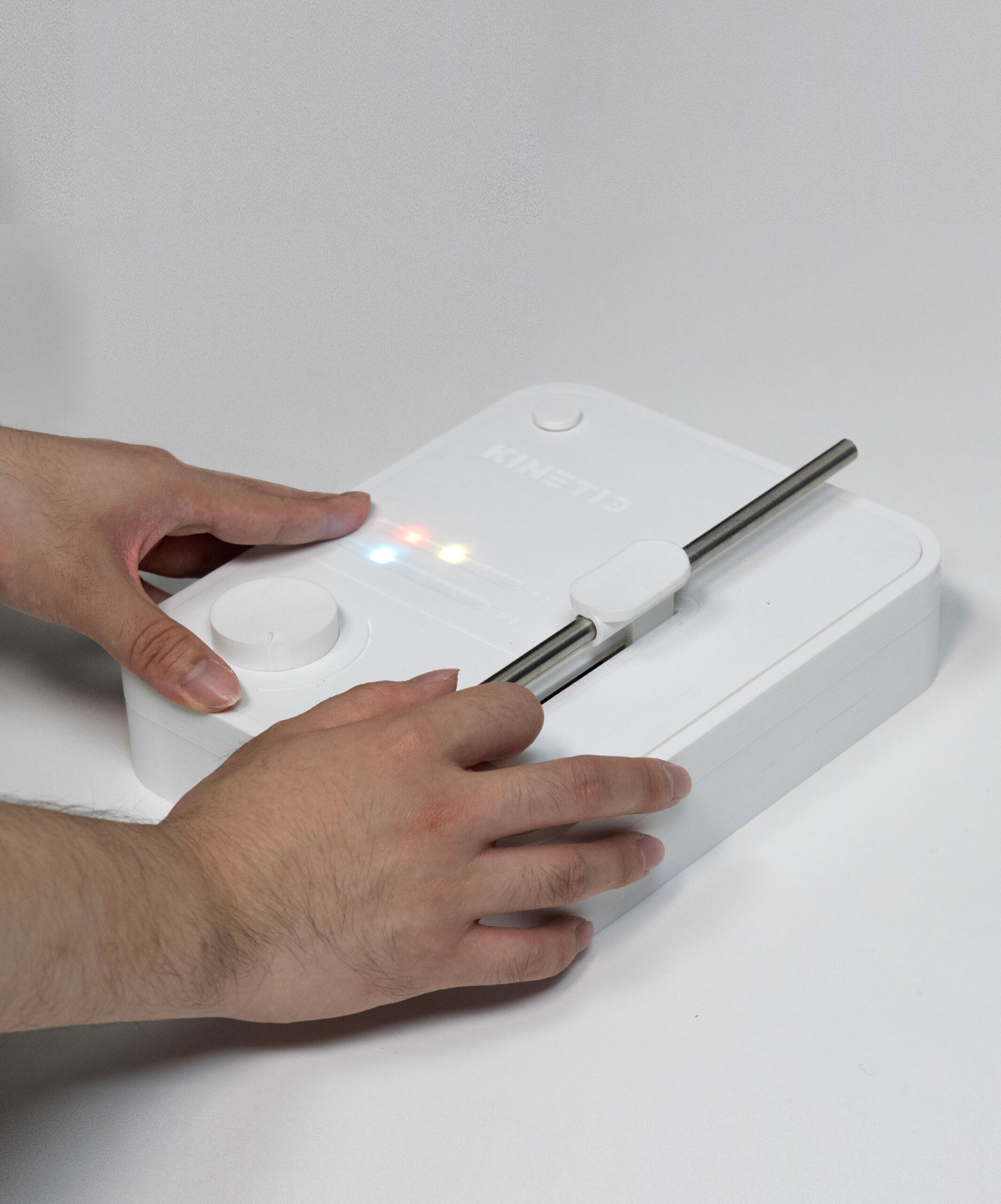 Two hands operating a white box-shaped device with diferent colored LED lights, an rotating knob, and a metallic rod.